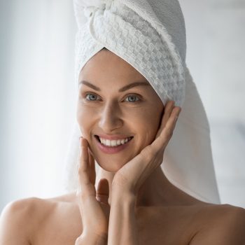 After shower body and head wrapped in towel 35s woman looks in mirror touches moisturized soft healthy face skin feels satisfied enjoy spa cosmetics treatment procedure, morning care hygiene concept